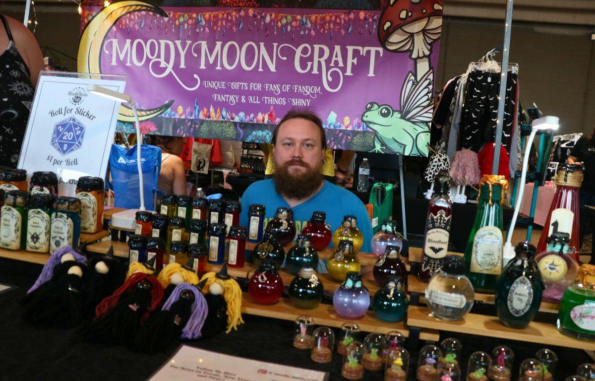 A man stands behind a colorful booth labeled "Moody Moon Craft," featuring fantasy-themed items like potion bottles, tiny figurines, and yarn witches. The setup includes a whimsical purple banner and a sign offering "$1 Roll for Sticker."