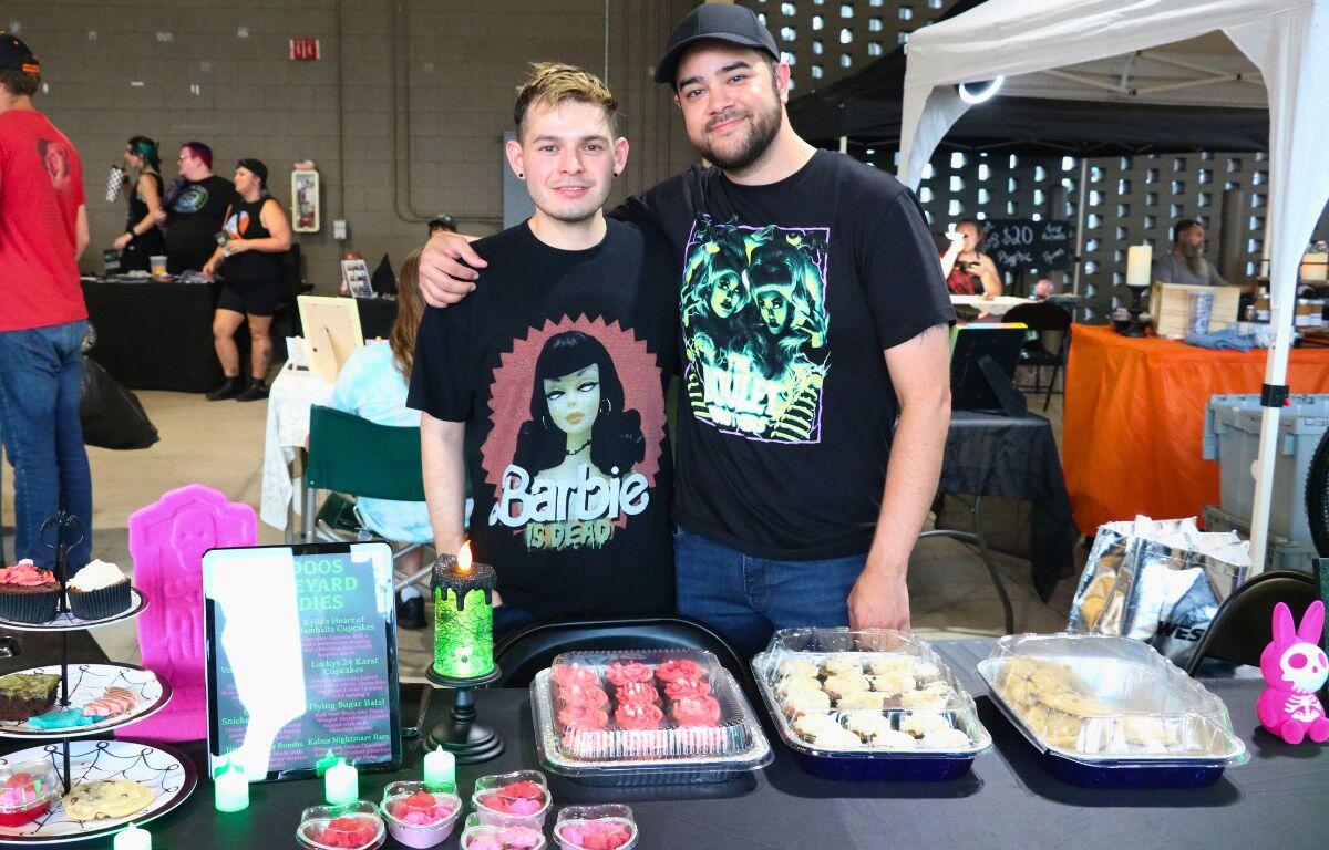Two men smile behind Halloween-themed baked goods at a canopied market featuring cupcakes, heart-shaped treats and spooky decorations.