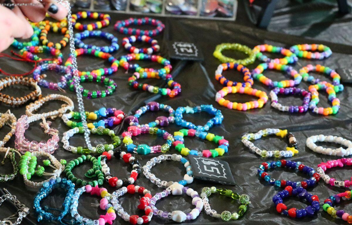 Multiple handmade bracelets are displayed on a table with a black tablecloth with signs reading, "$2" and "$5". Two hands lift a necklace over the bracelets.