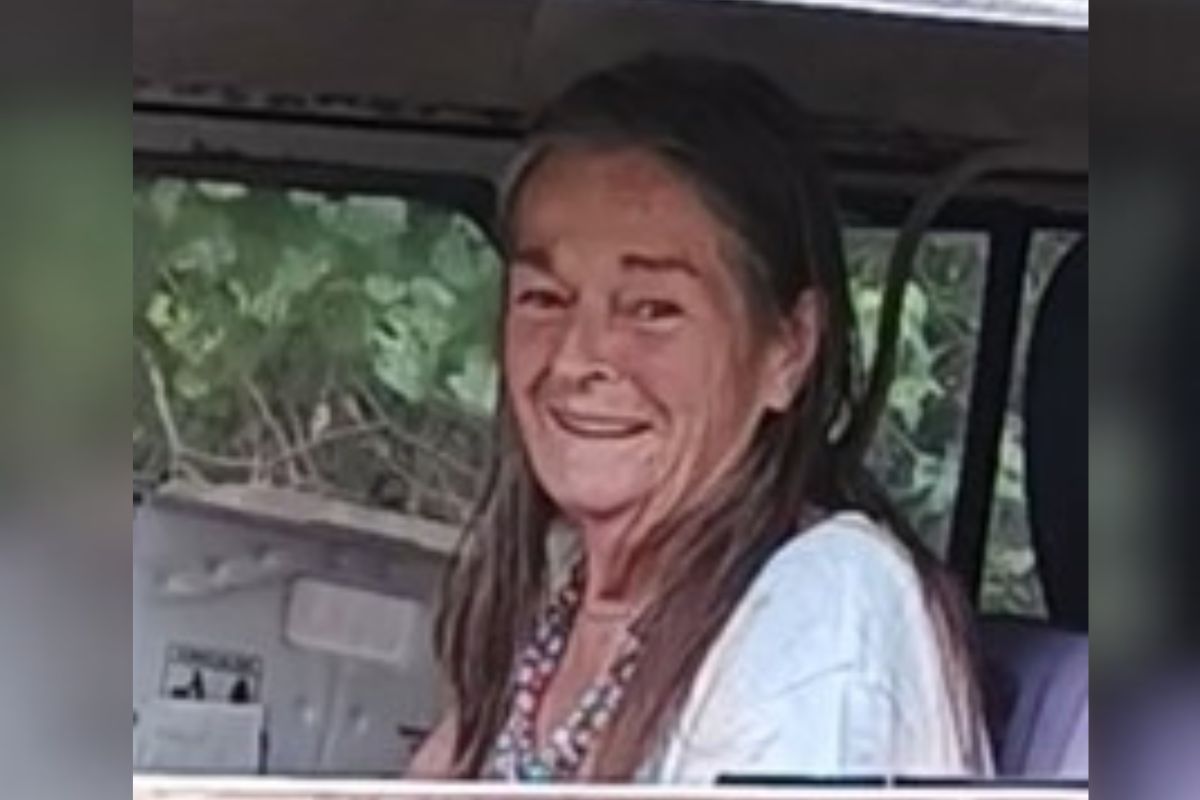 A woman sitting in the interior of a car, with flora, in the background.