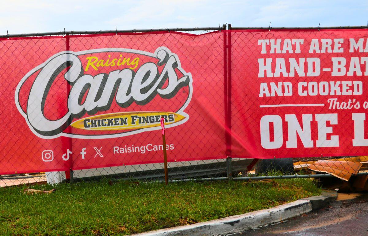 A red banner on a chain-link fence reads, "Raising Cane's Chicken Fingers" with social media logos beneath it. Next to the logos reads, "Raising Canes." In fron of the fence is a road and a patch of green grass next to it. Through the fence, construction materials can be seen.