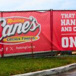 A red banner on a chain-link fence reads, "Raising Cane's Chicken Fingers" with social media logos beneath it. Next to the logos reads, "Raising Canes." In fron of the fence is a road and a patch of green grass next to it. Through the fence, construction materials can be seen.