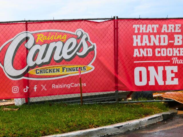 A red banner on a chain-link fence reads, "Raising Cane's Chicken Fingers" with social media logos beneath it. Next to the logos reads, "Raising Canes." In fron of the fence is a road and a patch of green grass next to it. Through the fence, construction materials can be seen.