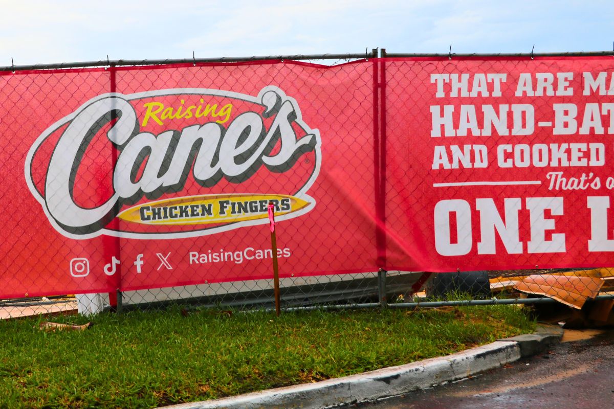 A red banner on a chain-link fence reads, "Raising Cane's Chicken Fingers" with social media logos beneath it. Next to the logos reads, "Raising Canes." In fron of the fence is a road and a patch of green grass next to it. Through the fence, construction materials can be seen.
