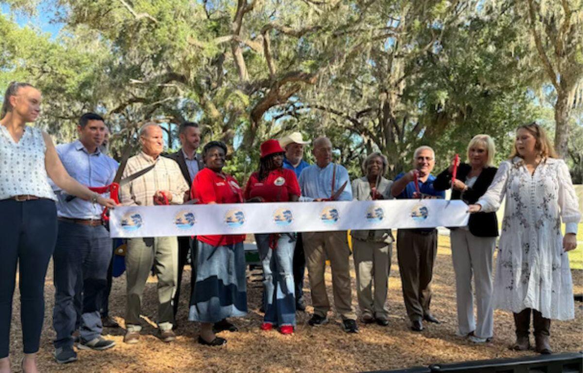 People standing behind a ribbon, with two people holding giant scissors to cut the ribbon. Trees in the background.