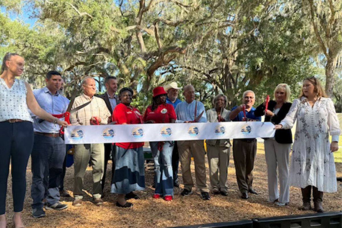 People standing behind a ribbon, with two people holding giant scissors to cut the ribbon. Trees in the background.