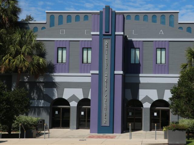 Front view of the Reilly Arts Center, featuring a gray and purple art deco facade with arched doorways and palm trees.