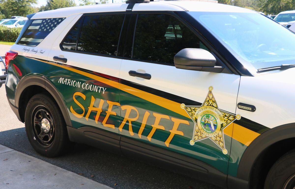 A front angle of a car with a green, yellow and white wrap with a monochrome American Flag on the back window sits next to a sidewalk. On the car reads, "Marion County Sheriff. Integrity. Honesty. Trust."