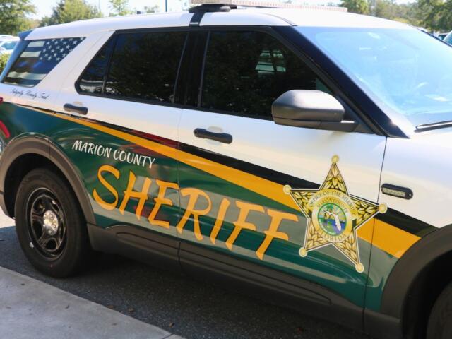 A front angle of a car with a green, yellow and white wrap with a monochrome American Flag on the back window sits next to a sidewalk. On the car reads, "Marion County Sheriff. Integrity. Honesty. Trust."