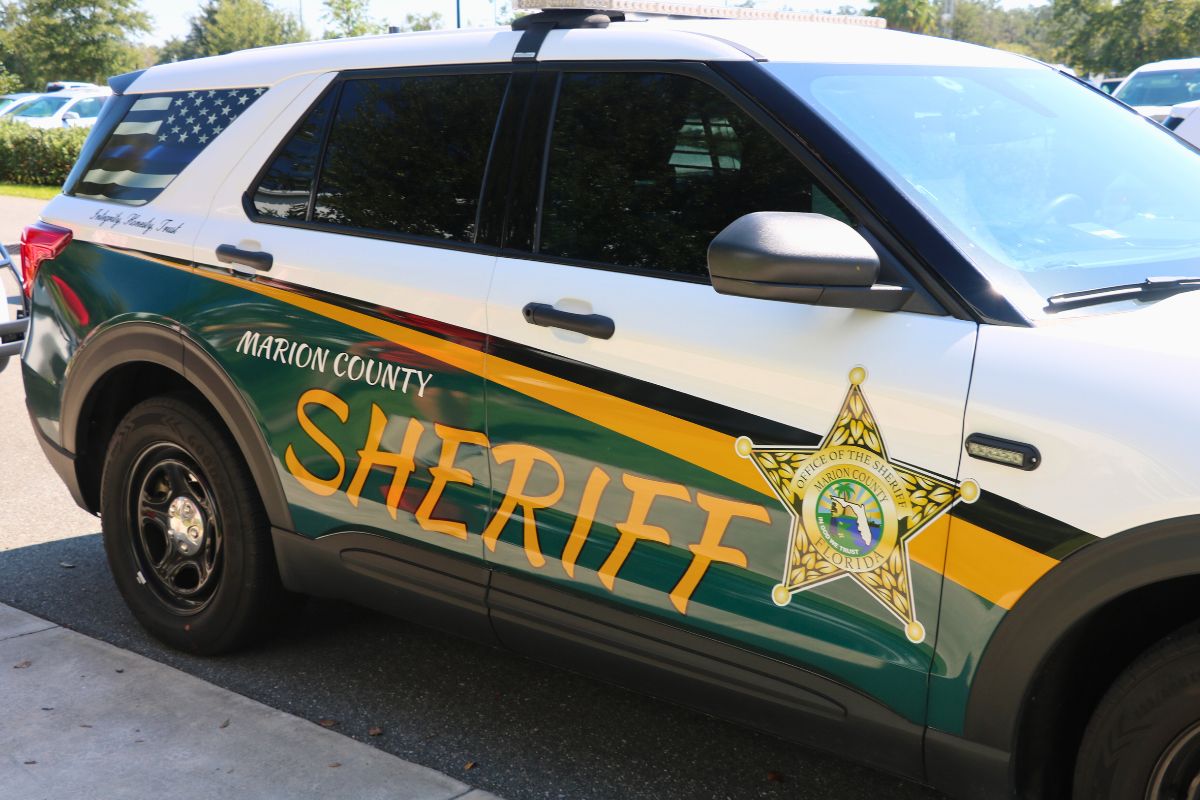 A front angle of a car with a green, yellow and white wrap with a monochrome American Flag on the back window sits next to a sidewalk. On the car reads, "Marion County Sheriff. Integrity. Honesty. Trust."