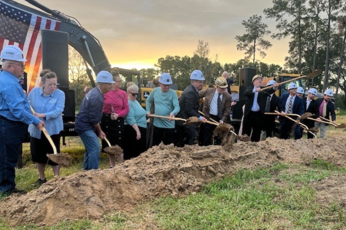 A group of people in hardhats, holding shovels with dirt, with the U.S.A. flag in the background, heavy equipment, the sunset and a tree line. In the foreground, there is a large pile of dirt and grass.