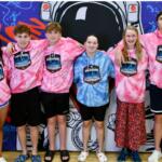 A group of young athletes, dressed in pink sweatshirts, with the exception of the one in the middle in a blue sweatshirt, standing in front of a colorful mural.