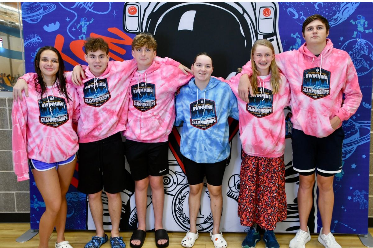 A group of young athletes, dressed in pink sweatshirts, with the exception of the one in the middle in a blue sweatshirt, standing in front of a colorful mural.