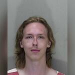 Mugshot of a young man with long, light brown hair wearing a red and white striped jail uniform, standing against a gray wall.