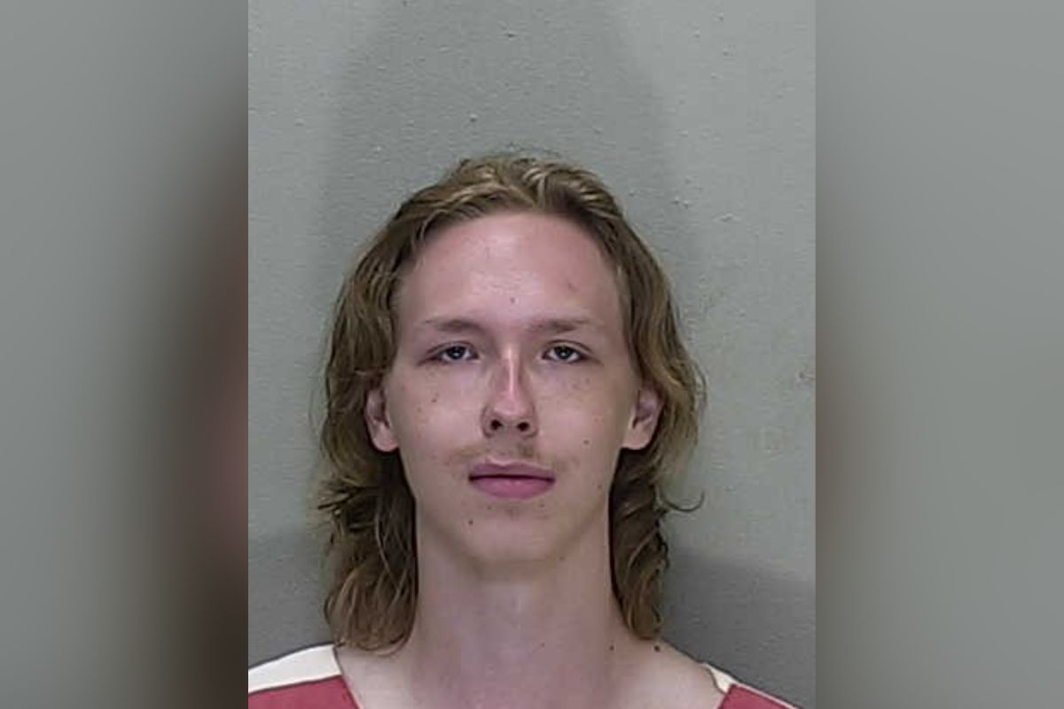 Mugshot of a young man with long, light brown hair wearing a red and white striped jail uniform, standing against a gray wall.