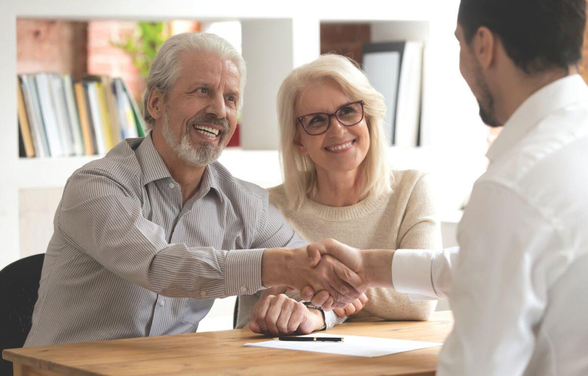 A smiling older couple sits across a desk from a professional, with the man shaking hands in agreement while the woman looks on approvingly.