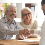A smiling older couple sits across a desk from a professional, with the man shaking hands in agreement while the woman looks on approvingly.