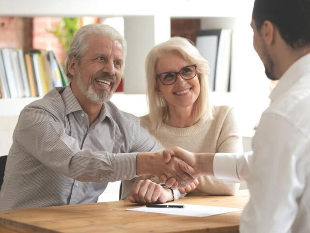 A smiling older couple sits across a desk from a professional, with the man shaking hands in agreement while the woman looks on approvingly.