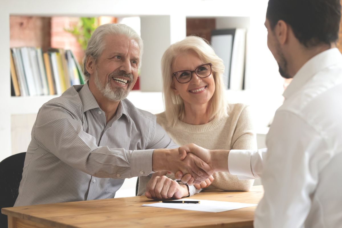 A smiling older couple sits across a desk from a professional, with the man shaking hands in agreement while the woman looks on approvingly.