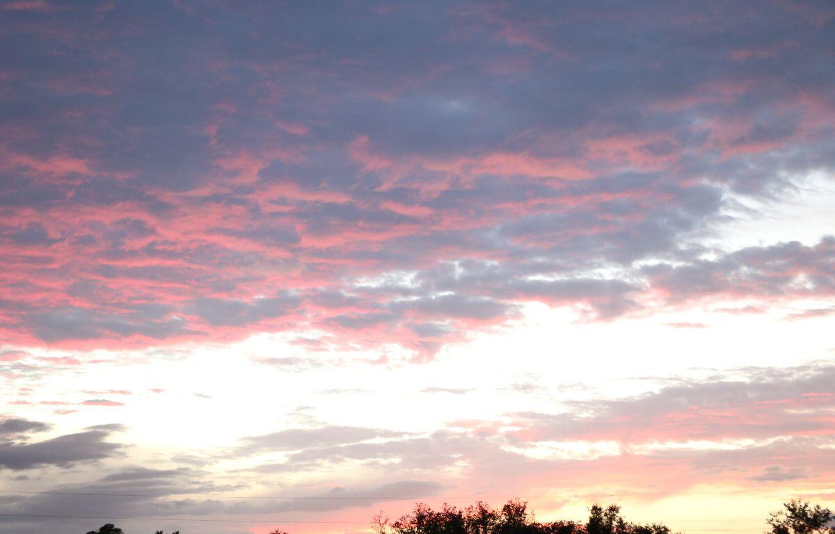 Dark clouds under a bright, white sky with dark pink and Amber hues underneath them, illuminated by a sunset hidden by a dark tree line toward the bottom.