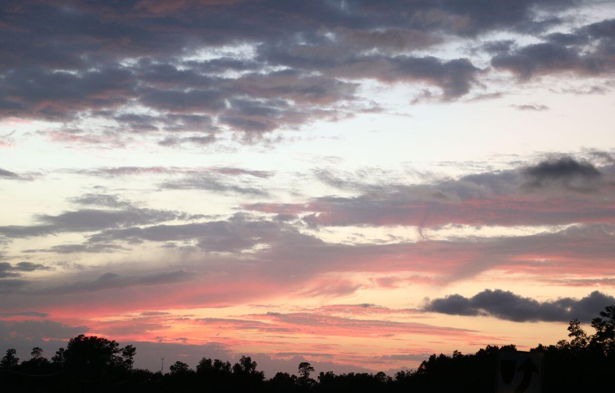 A sunset hidden behind a long, dark tree line provides the sky a final bit of light, making some clouds appear a dark pink and the sky an Amber color. Clouds further away are scattered and a cool blue and grey tone.