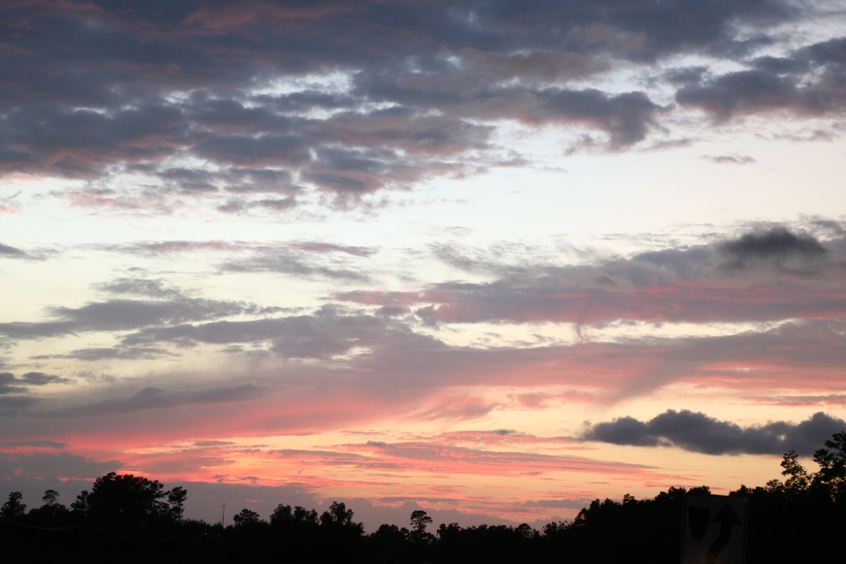 A sunset hidden behind a long, dark tree line provides the sky a final bit of light, making some clouds appear a dark pink and the sky an Amber color. Clouds further away are scattered and a cool blue and grey tone.
