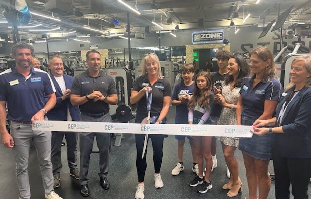 A group of people standing behind a ribbon, with the woman in the middle holding a giant pair of scissors, with exercise equipment and lighting in the background.