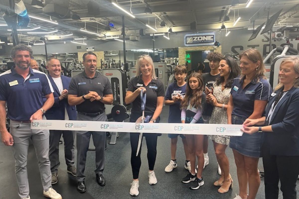 A group of people standing behind a ribbon, with the woman in the middle holding a giant pair of scissors, with exercise equipment and lighting in the background.