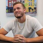 Country artist Noah Hunton smiles while seated at a table during an interview, wearing a white shirt and gold chain with a mural of downtown buildings in the background.