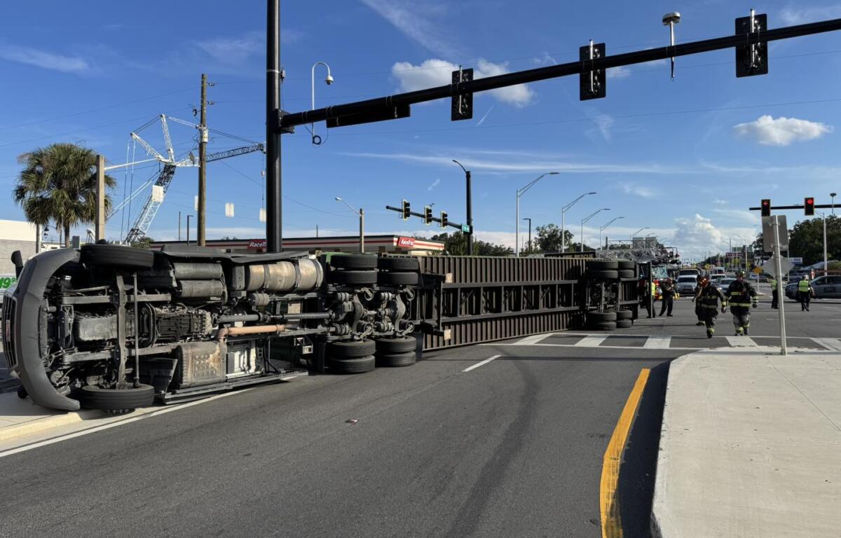 A large semi-truck is overturned on its side, blocking multiple lanes of a busy intersection under a clear blue sky. Emergency responders, including firefighters, are on the scene managing the situation.
