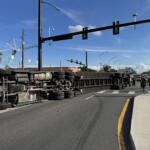 A large semi-truck is overturned on its side, blocking multiple lanes of a busy intersection under a clear blue sky. Emergency responders, including firefighters, are on the scene managing the situation.
