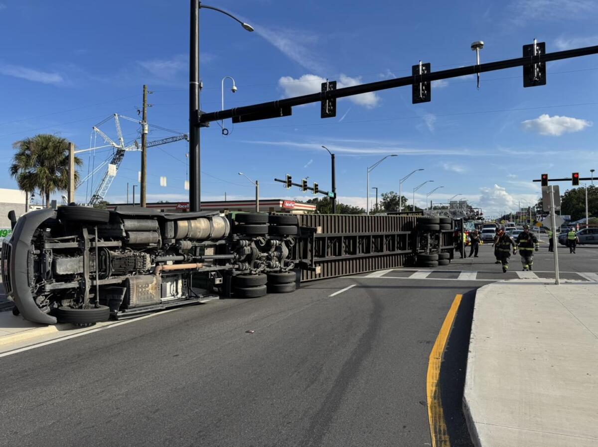 A large semi-truck is overturned on its side, blocking multiple lanes of a busy intersection under a clear blue sky. Emergency responders, including firefighters, are on the scene managing the situation.