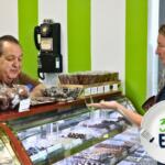 A man stands behind a display counter with a variety of chocolate items on display in the case and on top of it, a vase of purple flowers and an old-fashion phone on a green and white wall behind the man. A girl stands on the other side of the counter with a chocolate truffle in her hand.