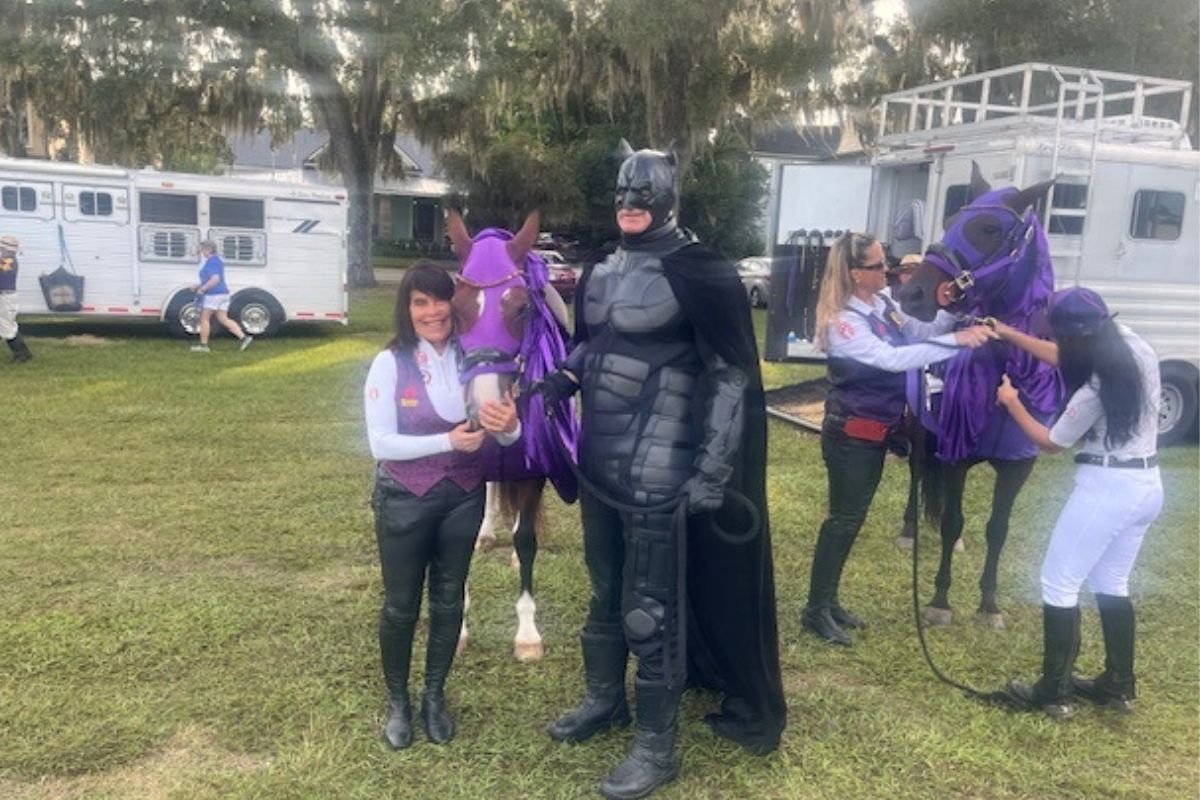 To the left, horse trailers in the background, in the center a woman in decorative riding attire, a mare and a superhero figure, to the right in the background a horse trailer and truck with two women with a horse with a trailer directly behind them.