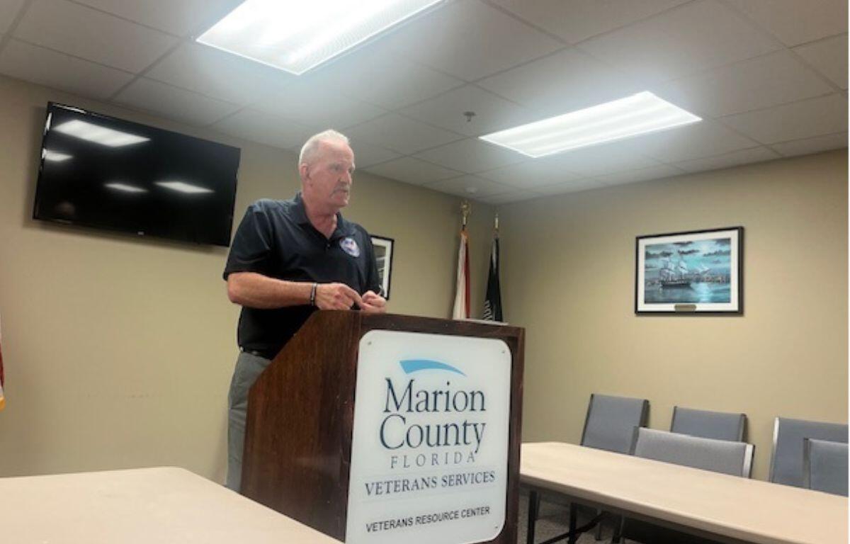 TV monitor mounted to the backwall, a man in a blue shirt behind a podium with a microphone, with a white placard with writing and an insignia on it. A table. to the left, and a table with chairs to the right and a picture on the wall.