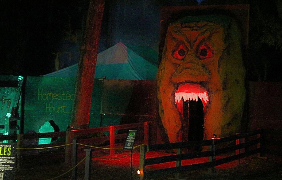 A large alien head facade stands tall in front of a building with an open mouth for guests to walk through. There are canopies in the background with writing on a wall reading, "Homestead Haunt."