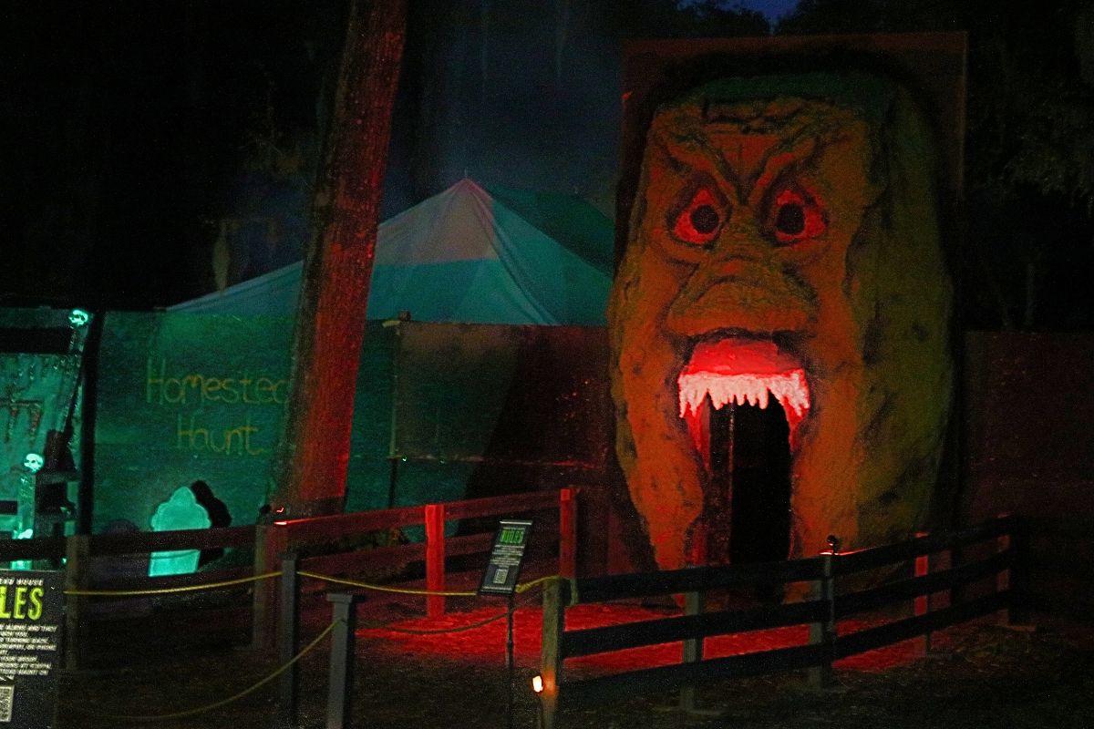A large alien head facade stands tall in front of a building with an open mouth for guests to walk through. There are canopies in the background with writing on a wall reading, "Homestead Haunt."