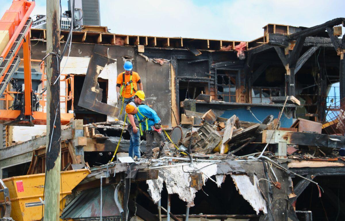 Construction workers in safety gear stand among debris and burned wooden beams of a partially destroyed building.