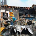 Construction workers in safety gear stand among debris and burned wooden beams of a partially destroyed building.
