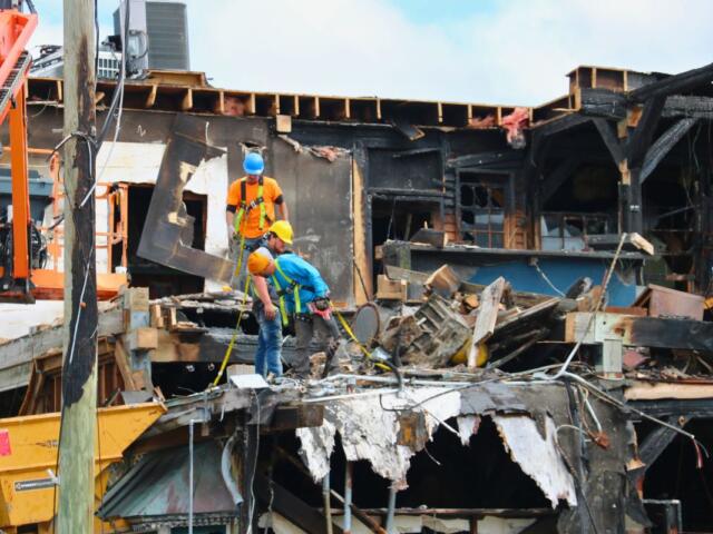 Construction workers in safety gear stand among debris and burned wooden beams of a partially destroyed building.
