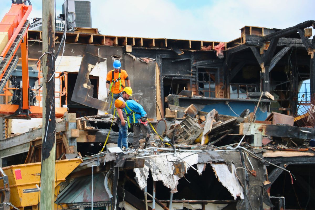 Construction workers in safety gear stand among debris and burned wooden beams of a partially destroyed building.