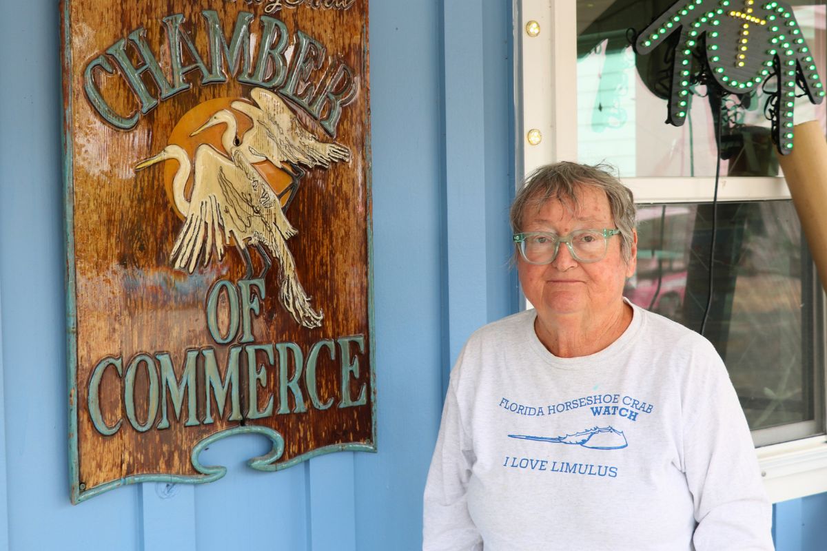 A woman wearing glasses and a “Florida Horseshoe Crab Watch” shirt stands beside a wooden “Chamber of Commerce” sign on a blue wall.