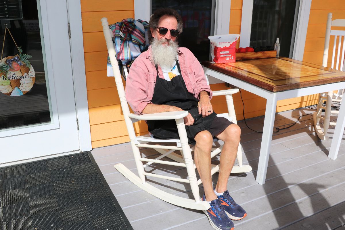 A man with a white beard and sunglasses sits in a white rocking chair on a porch painted bright yellow. He wears a pink shirt, black apron, and blue sneakers.