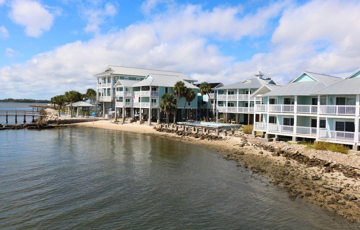 A shoreline leads to a sandy and rocky area with a pier leading out into the water. Lifted blue building with gray roofs and white railings line the shore.