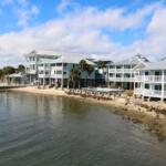 A shoreline leads to a sandy and rocky area with a pier leading out into the water. Lifted blue building with gray roofs and white railings line the shore.