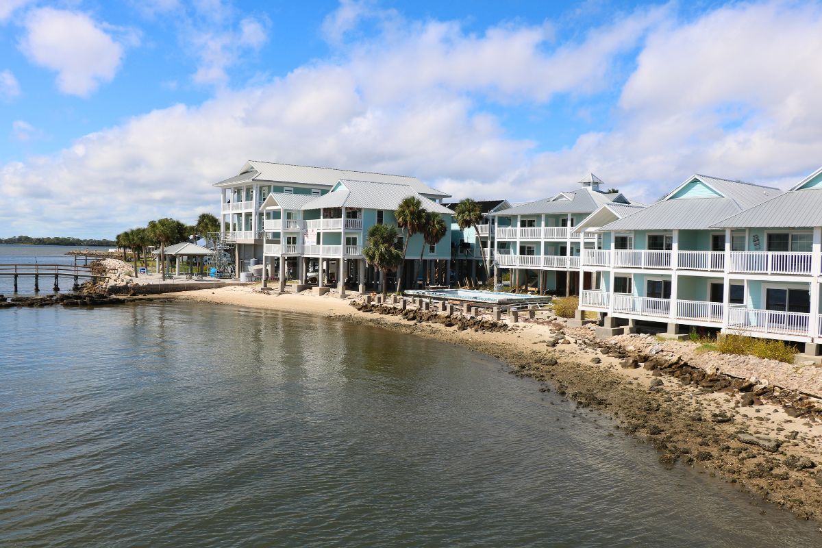 A shoreline leads to a sandy and rocky area with a pier leading out into the water. Lifted blue building with gray roofs and white railings line the shore.