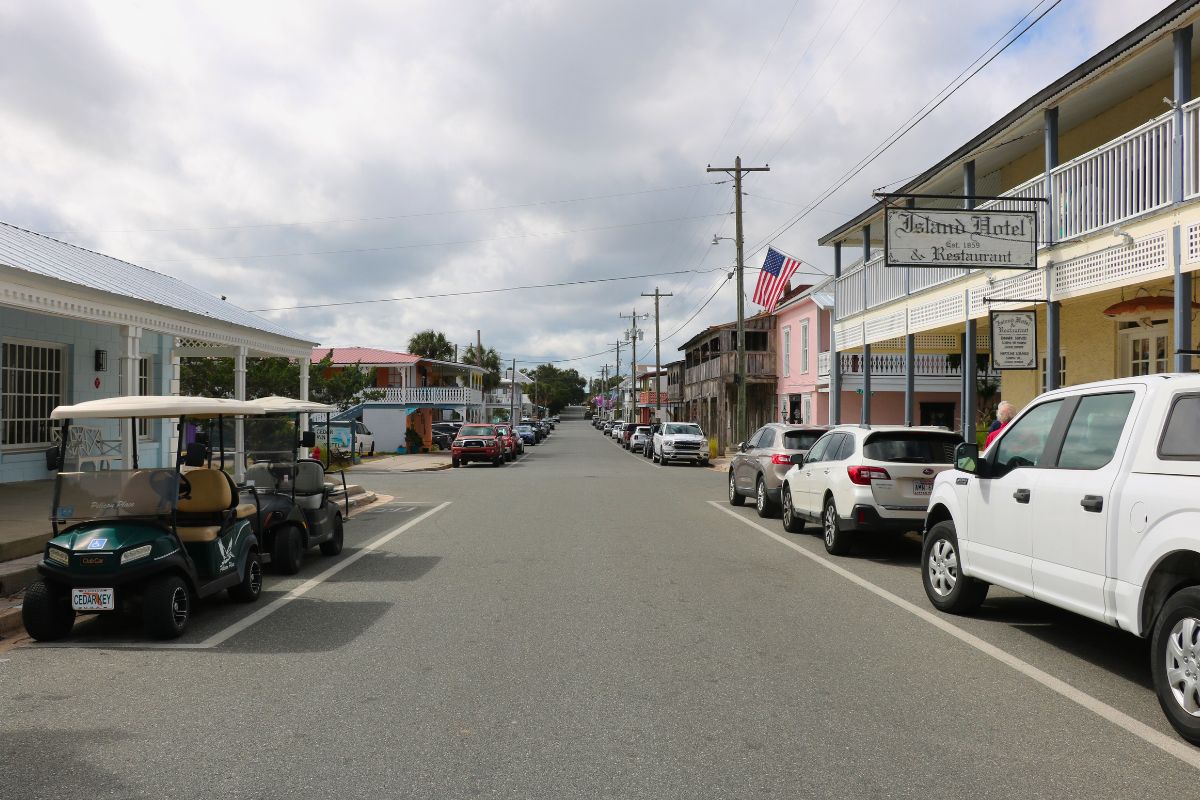 A street is line with cars and golf carts in parallel parking spots with buildings on either side of the road leading into the distance where the street continues to have trees on either side.