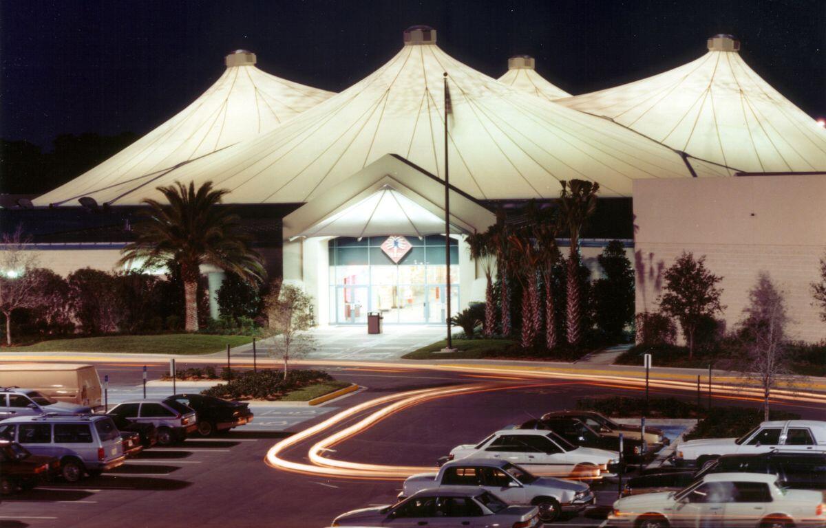 An enclosed mall with a white tent canopy covering and brick walls. Palm trees decorate the entrance with antique cars in the parking lot leaving for the day. The white tent is illuminated against the night sky.