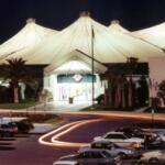An enclosed mall with a white tent canopy covering and brick walls. Palm trees decorate the entrance with antique cars in the parking lot leaving for the day. The white tent is illuminated against the night sky.