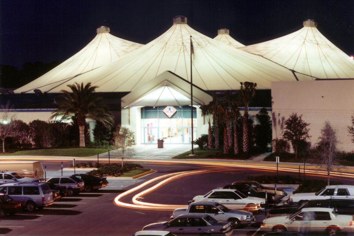 An enclosed mall with a white tent canopy covering and brick walls. Palm trees decorate the entrance with antique cars in the parking lot leaving for the day. The white tent is illuminated against the night sky.